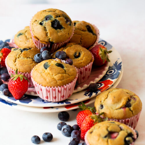 Un plato de muffins de frutos rojos recién horneados sobre una vajilla decorativa. Los muffins, en moldes de rayas rosas, rebosan de arándanos. A su alrededor, frutos rojos frescos como arándanos y fresas. La escena se desarrolla sobre un fondo suave y claro, creando un cuadro encantador.