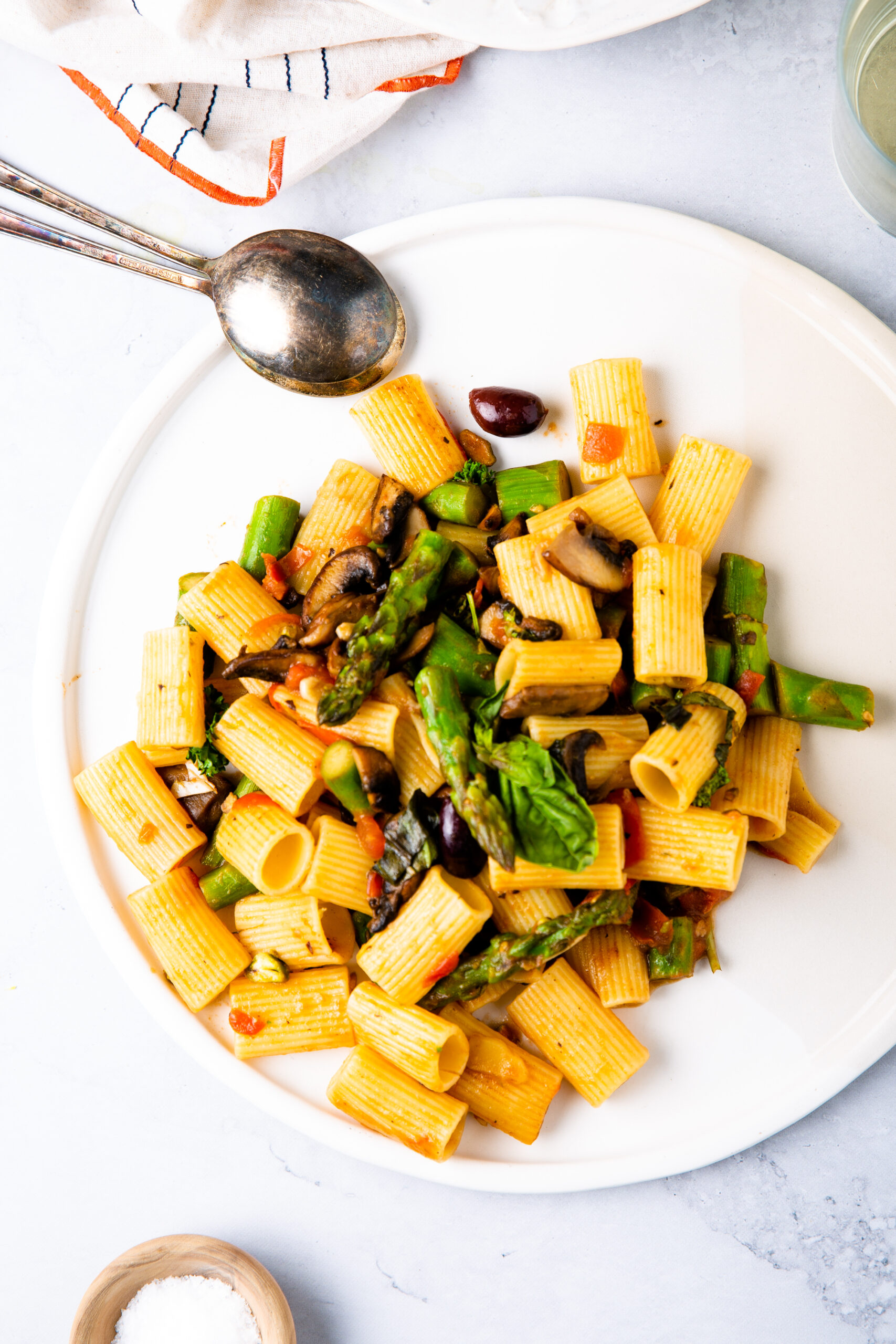 Un plato blanco lleno de pasta rigatoni mezclada con espárragos verdes, espinacas, champiñones, tomates y aceitunas negras. Una cuchara y un tenedor de metal descansan sobre el plato, con un tazón de sal gruesa y una servilleta de tela visibles cerca sobre una superficie clara.