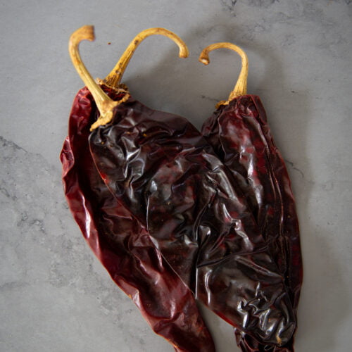 Dos chiles rojos secos con cáscaras arrugadas y brillantes y tallos curvos de color canela descansan sobre una superficie gris claro, ligeramente jaspeada, perfecta para añadir un picante ahumado a platos como la sopa de frijol o para servir junto a nopales y crujientes totopos.