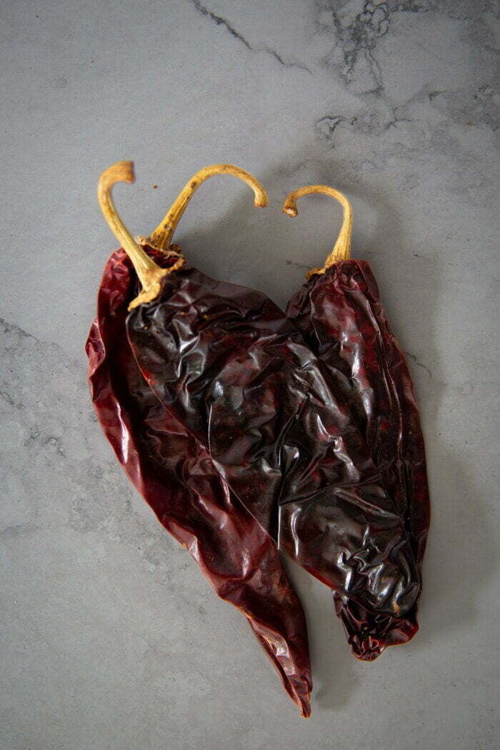 Dos chiles rojos secos con c&aacute;scaras arrugadas y brillantes y tallos curvos de color canela descansan sobre una superficie gris claro, ligeramente jaspeada, perfecta para a&ntilde;adir un picante ahumado a platos como la sopa de frijol o para servir junto a nopales y crujientes totopos.