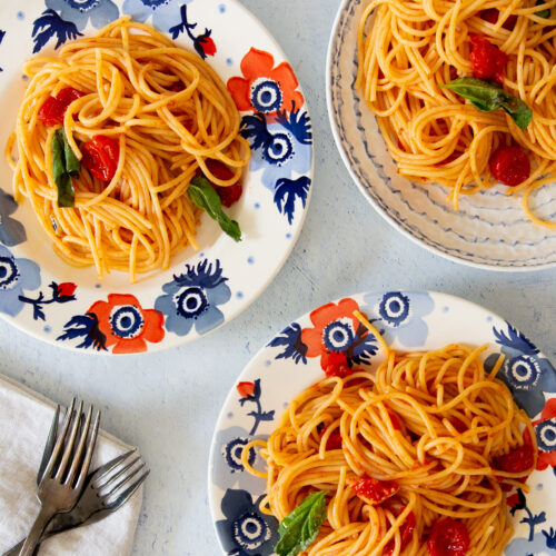 Tres platos de pasta con tomates cherry y albahaca, dispuestos sobre una superficie azul claro.