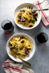 Sobre una superficie de color claro se encuentran dos cuencos blancos de pasta con champiñones y hierbas. Cada cuenco tiene un tenedor, uno dorado y otro plateado. Junto a los cuencos hay dos copas llenas de vino tinto. Junto a cada cuenco se coloca una servilleta de rayas blancas y rojas, que añade un toque de elegancia a la escena bien iluminada.