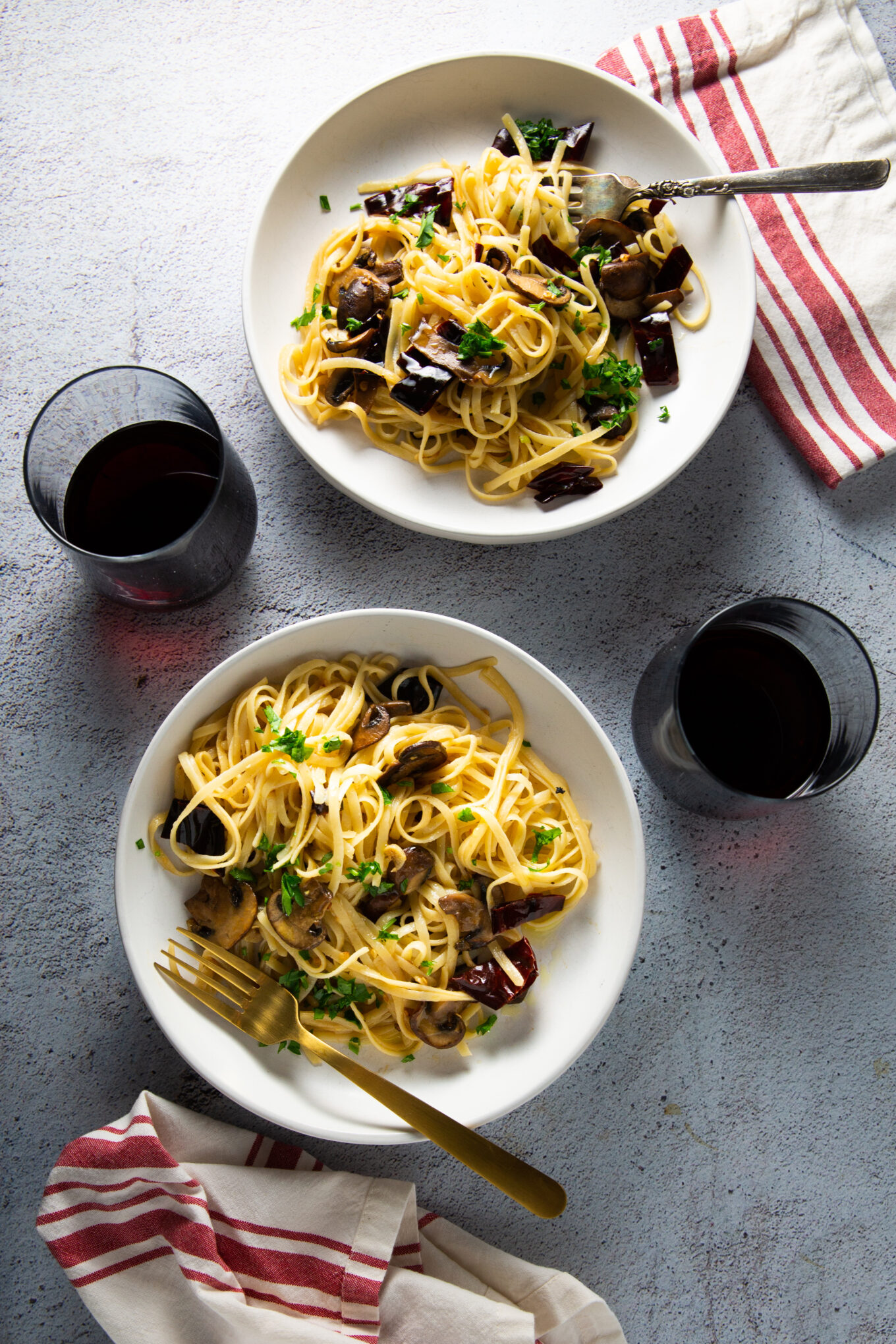 Sobre una superficie de color claro se encuentran dos cuencos blancos de pasta con champiñones y hierbas. Cada cuenco tiene un tenedor, uno dorado y otro plateado. Junto a los cuencos hay dos copas llenas de vino tinto. Junto a cada cuenco se coloca una servilleta de rayas blancas y rojas, que añade un toque de elegancia a la escena bien iluminada.