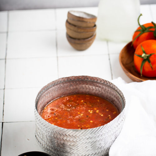 Un tazón de metal lleno de sopa de tomate espesa reposa sobre una superficie de azulejos blancos. Cerca, se ve una cuchara de madera con un poco de sopa y una pequeña tapa negra. Al fondo, junto al tazón de madera con tomates y otros tazones marrones apilados, yace una botella con corcho. Un mantel blanco cubre elegantemente esta vibrante escena, que evoca la preparación de una salsa mexicana de dos ingredientes.