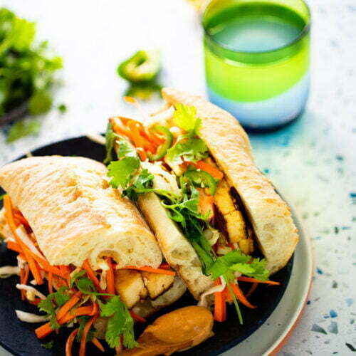 Un colorido sándwich con pollo a la parrilla, zanahorias ralladas, cilantro y jalapeños sobre rebanadas de baguette crujientes evoca la vibrante esencia de un bánh mì. Se coloca en un plato junto a una cuchara de madera llena de salsa. Cerca, un vaso con degradado verde y azul contiene agua, con hierbas y limón de fondo.
