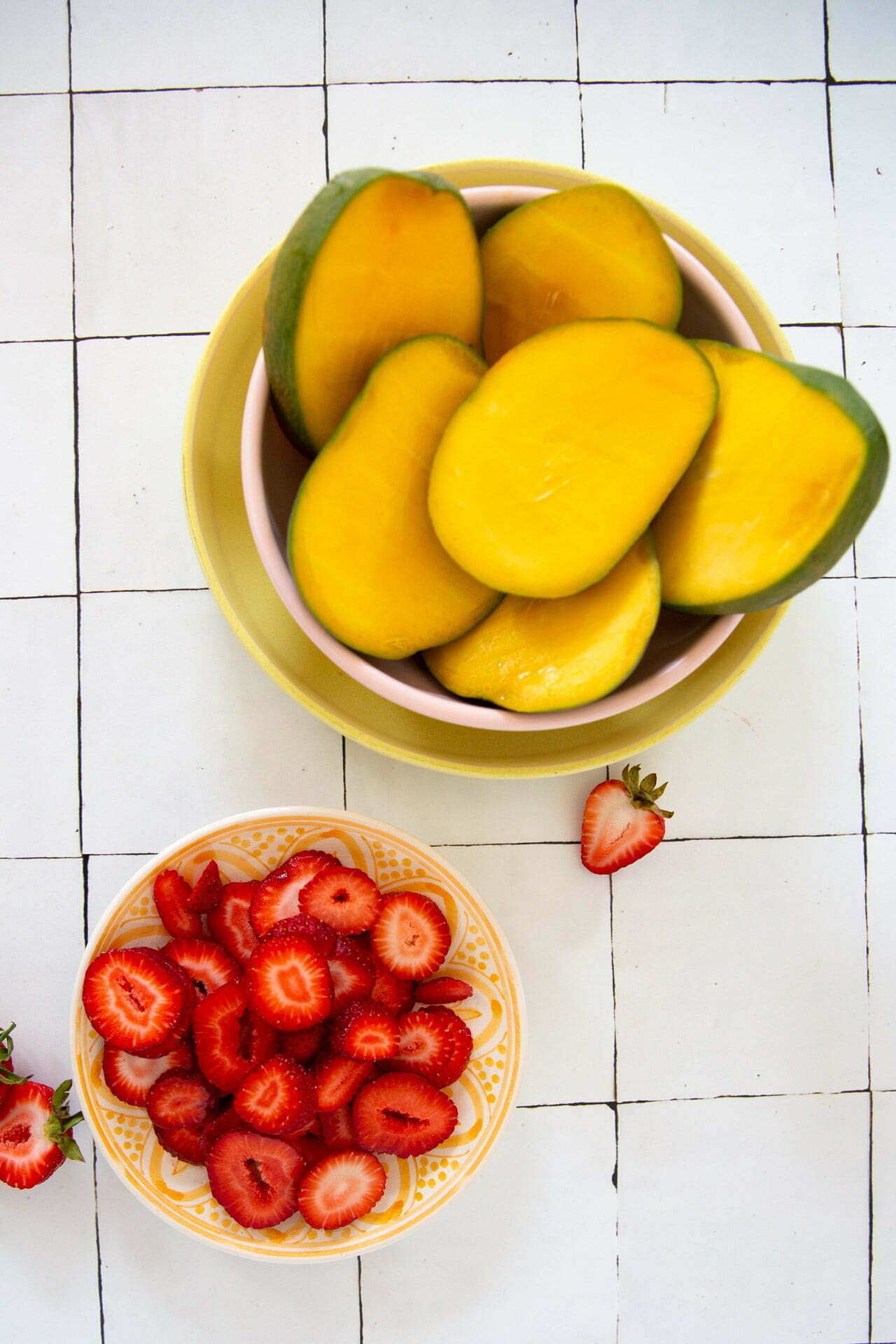 Tazones sobre una superficie de azulejos: uno con rodajas de mango que recuerdan a las paletas de mango y el otro rebosante de fresas rebanadas. Dos fresas enteras se colocan junto a los tazones como decoración. Las rodajas de mango de un amarillo vibrante y las fresas de un rojo intenso crean una presentación colorida y fresca.