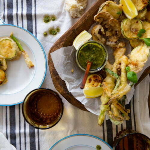 A spread of fried vegetables arranged in a wooden tray lined with parchment paper, accompanied by lemon wedges and a green dipping sauce. Two plates of vegetable fritto misto sit beside the tray, and two amber glasses are set nearby. A piece of baguette is visible in the top left corner.