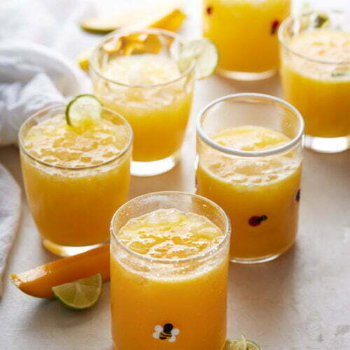 Seis vasos llenos de agua fresca de mango de color naranja están dispuestos sobre una superficie blanca. Las bebidas están adornadas con cubitos de hielo y algunas tienen rodajas de lima en el borde. Los vasos presentan pequeños estampados decorativos. Alrededor de los vasos hay rodajas de lima y pequeñas rodajas de naranja. A la izquierda se ve parcialmente un paño blanco.