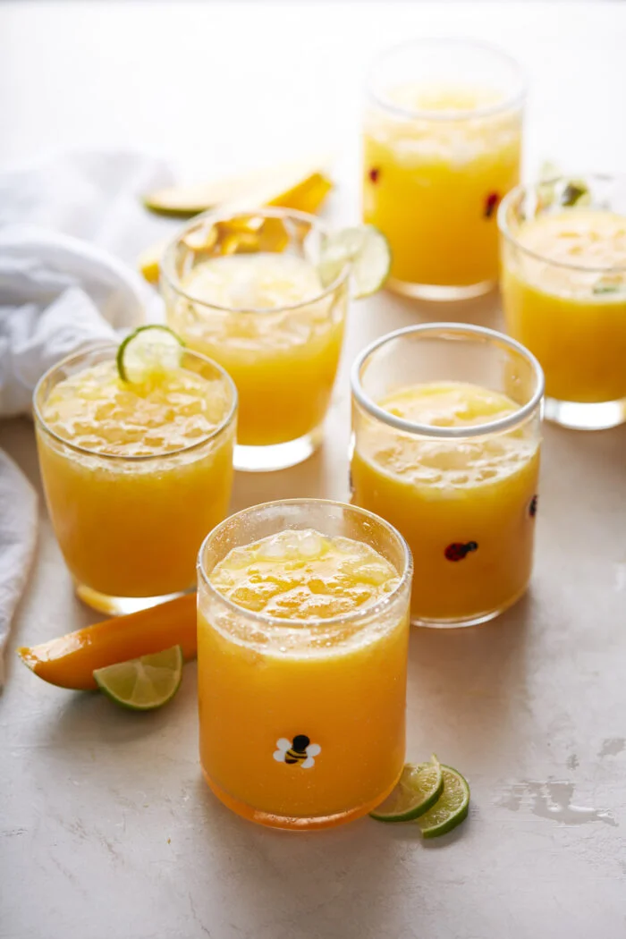 Seis vasos llenos de agua fresca de mango de color naranja est&aacute;n dispuestos sobre una superficie blanca. Las bebidas est&aacute;n adornadas con cubitos de hielo y algunas tienen rodajas de lima en el borde. Los vasos presentan peque&ntilde;os estampados decorativos. Alrededor de los vasos hay rodajas de lima y peque&ntilde;as rodajas de naranja. A la izquierda se ve parcialmente un pa&ntilde;o blanco.