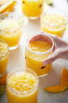 Una mano alcanza un vaso lleno de agua fresca de mango, rodeado por varios vasos similares sobre una superficie de color claro. Los vasos contienen cubitos de hielo y están adornados con rodajas de lima y trozos de mango. La escena es luminosa, con un aire fresco y veraniego.