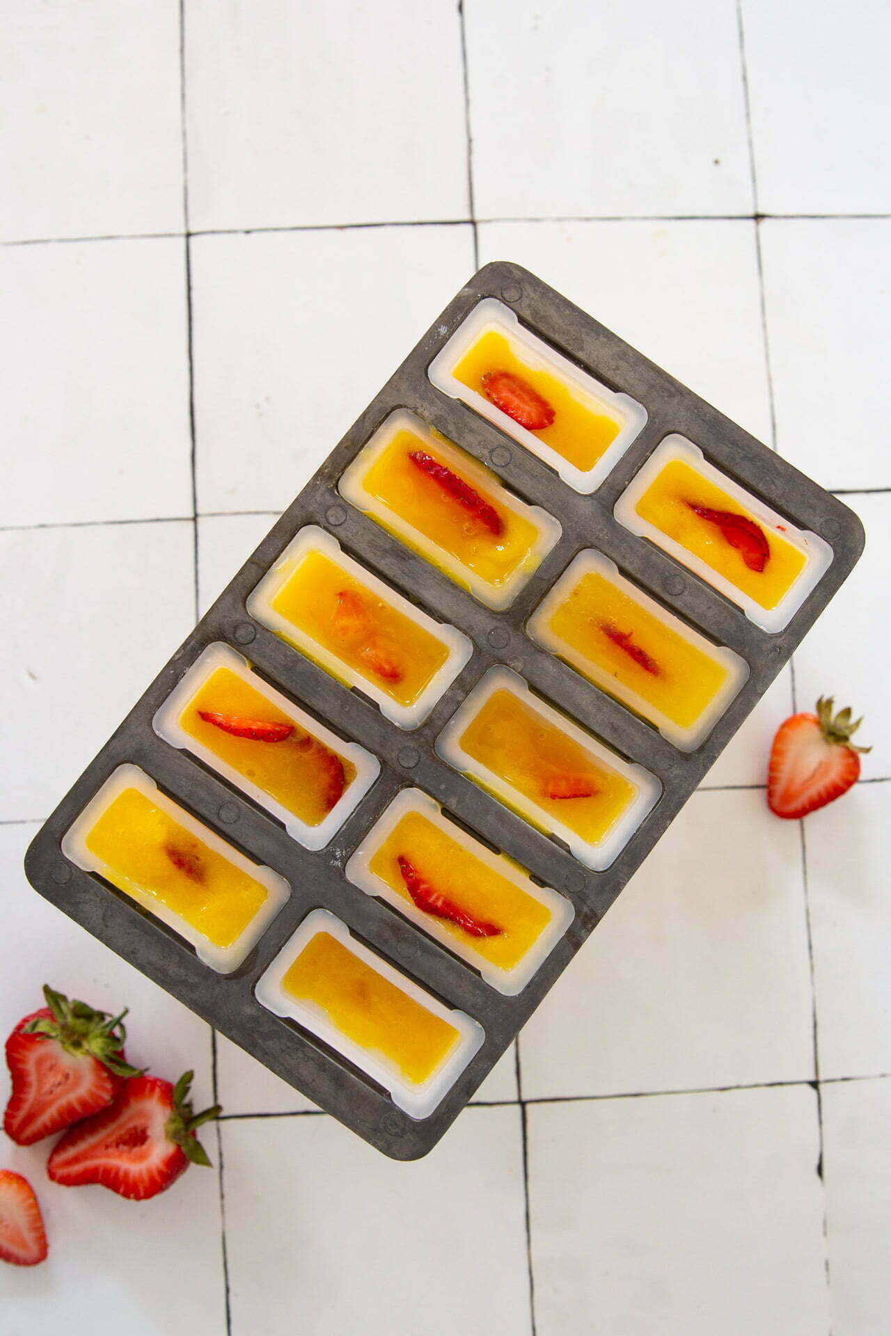 Un molde metálico para paletas, lleno de un líquido naranja que recuerda a las paletas de mango, con rodajas de fresa, se coloca sobre una superficie de baldosas blancas. Dos fresas frescas y algunas rebanadas descansan junto al molde, completando esta refrescante creación frutal.