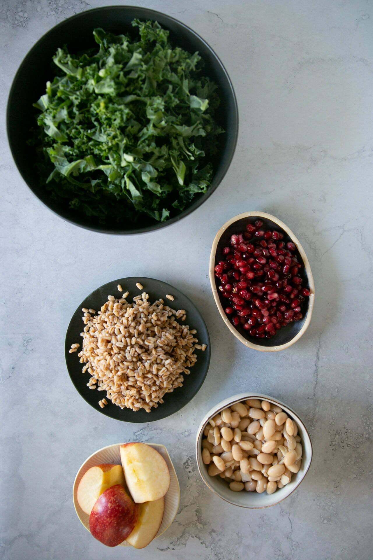 Cuatro cuencos y un plato muestran los frescos y coloridos ingredientes de una ensalada de farro: col rizada picada, granos de granada, farro cocido, judías blancas y manzanas rojas cortadas en rodajas dispuestas en forma circular sobre una superficie gris claro.