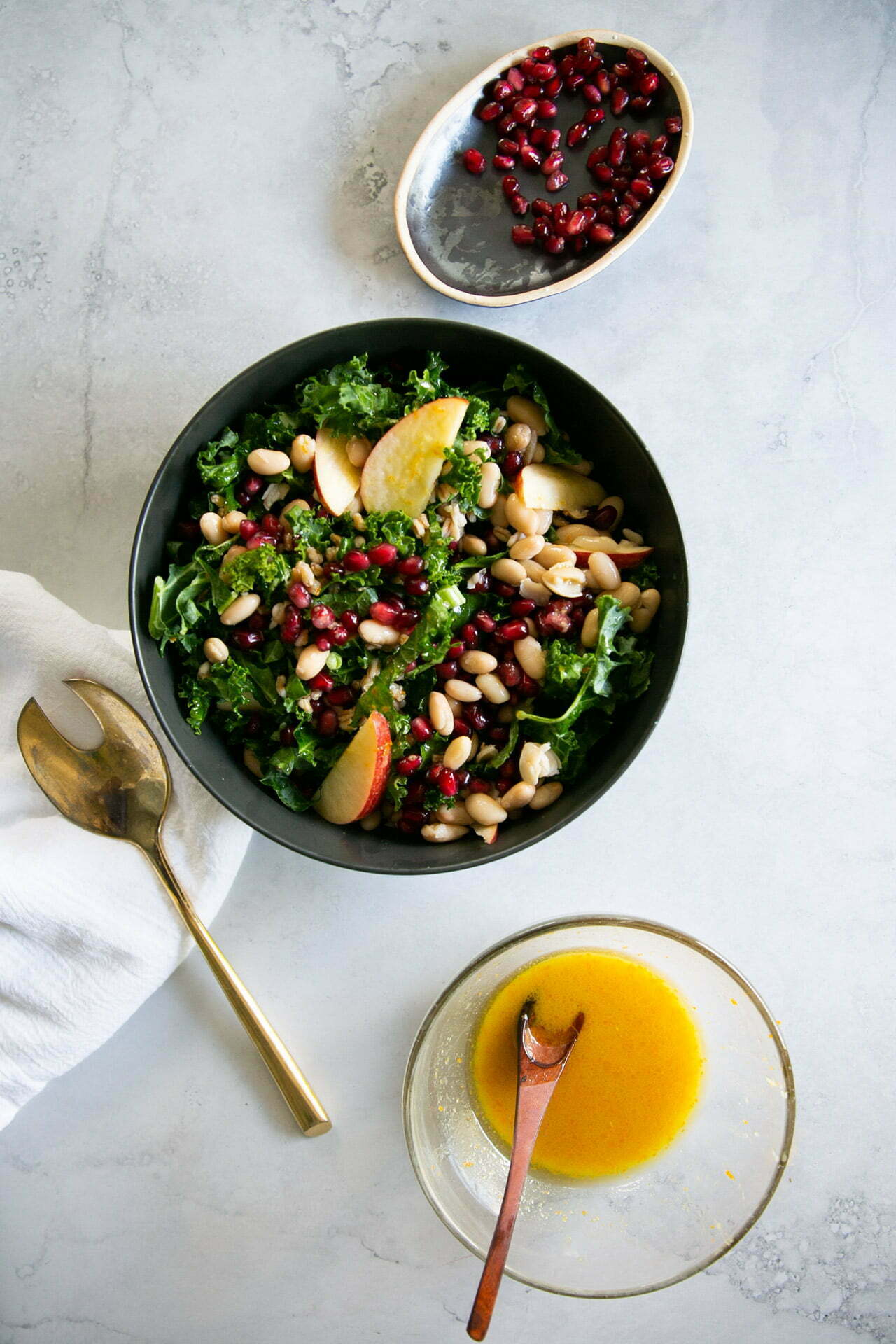 Un bol de ensalada con col rizada, rodajas de manzana, judías blancas y granos de granada descansa sobre una superficie clara. Cerca hay un tenedor de ensalada dorado, una servilleta blanca y un cuenco de aliño amarillo con una cuchara, perfecto para acompañar una ensalada de farro.