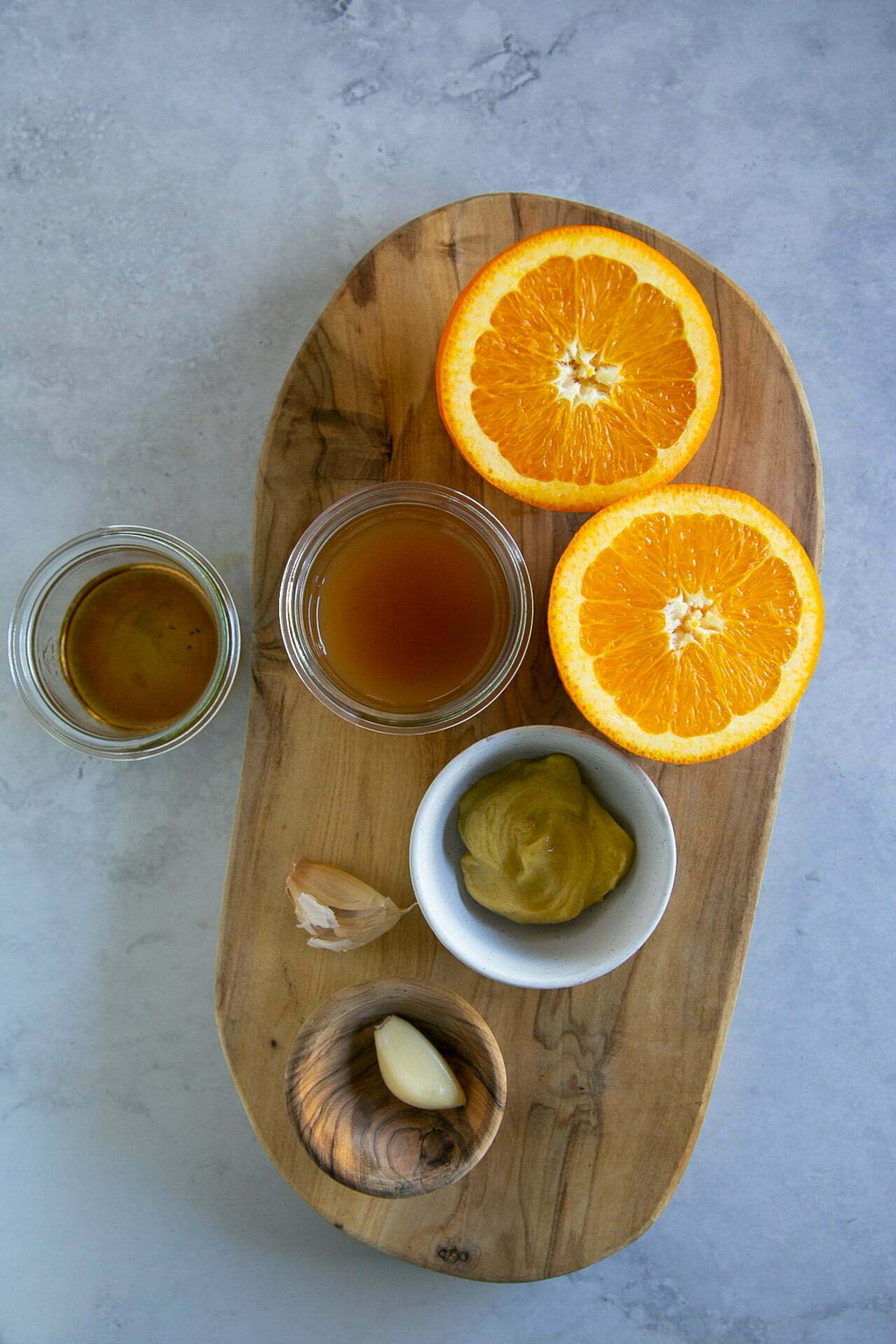 Una tabla de madera sostiene dos mitades de naranja, un pequeño cuenco de mostaza, dos dientes de ajo (uno pelado) y un tarro de líquido marrón, ingredientes perfectamente dispuestos para preparar una fresca ensalada de farro. Cerca hay otro tarro con líquido marrón dorado sobre una superficie gris claro.