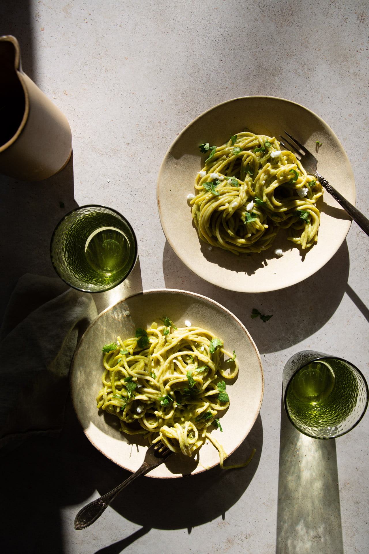 Dos platos de espagueti verde vegano cremosos con guarnición de hierbas están parcialmente expuestos a la luz del sol, lo que genera sombras. Hay tenedores sobre los platos. Dos vasos de vidrio verde acompañan la comida: uno vacío y genera sombra, el otro medio lleno. A la izquierda se ve parcialmente una jarra beige.