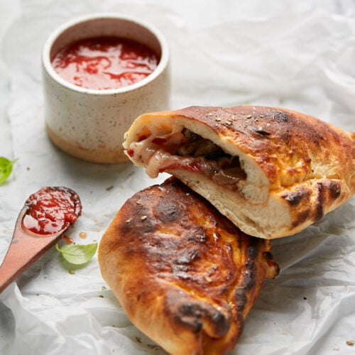 A golden-brown calzone, sliced open, reveals melted cheese and vegetables. It rests on crumpled white parchment paper beside a small ceramic bowl brimming with tomato sauce and a wooden spoon. A few basil leaves are artfully scattered around, adding a fresh touch to the savory scene.