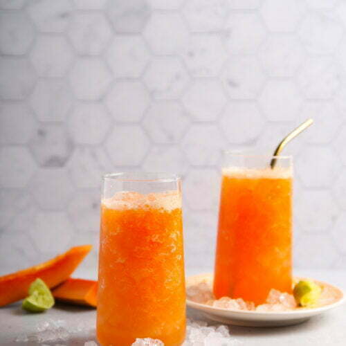Two tall glasses filled with bright orange papaya juice slush are on a light surface. One glass has a metal straw. A plate beside the glasses holds lime wedges and two papaya slices. Ice cubes are scattered around, set against a white hexagonal tile wall backdrop.