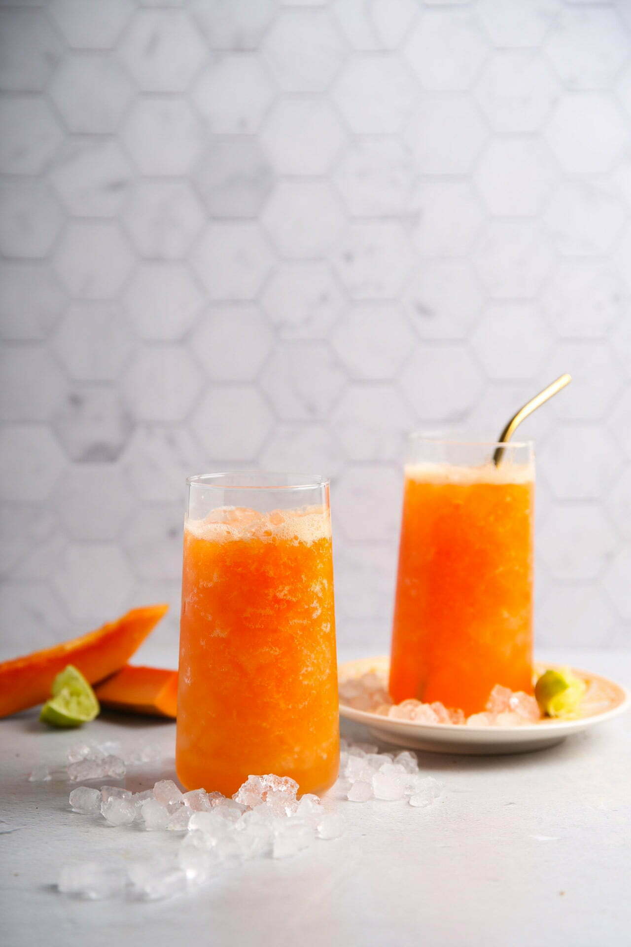Two tall glasses filled with bright orange papaya juice slush are on a light surface. One glass has a metal straw. A plate beside the glasses holds lime wedges and two papaya slices. Ice cubes are scattered around, set against a white hexagonal tile wall backdrop.
