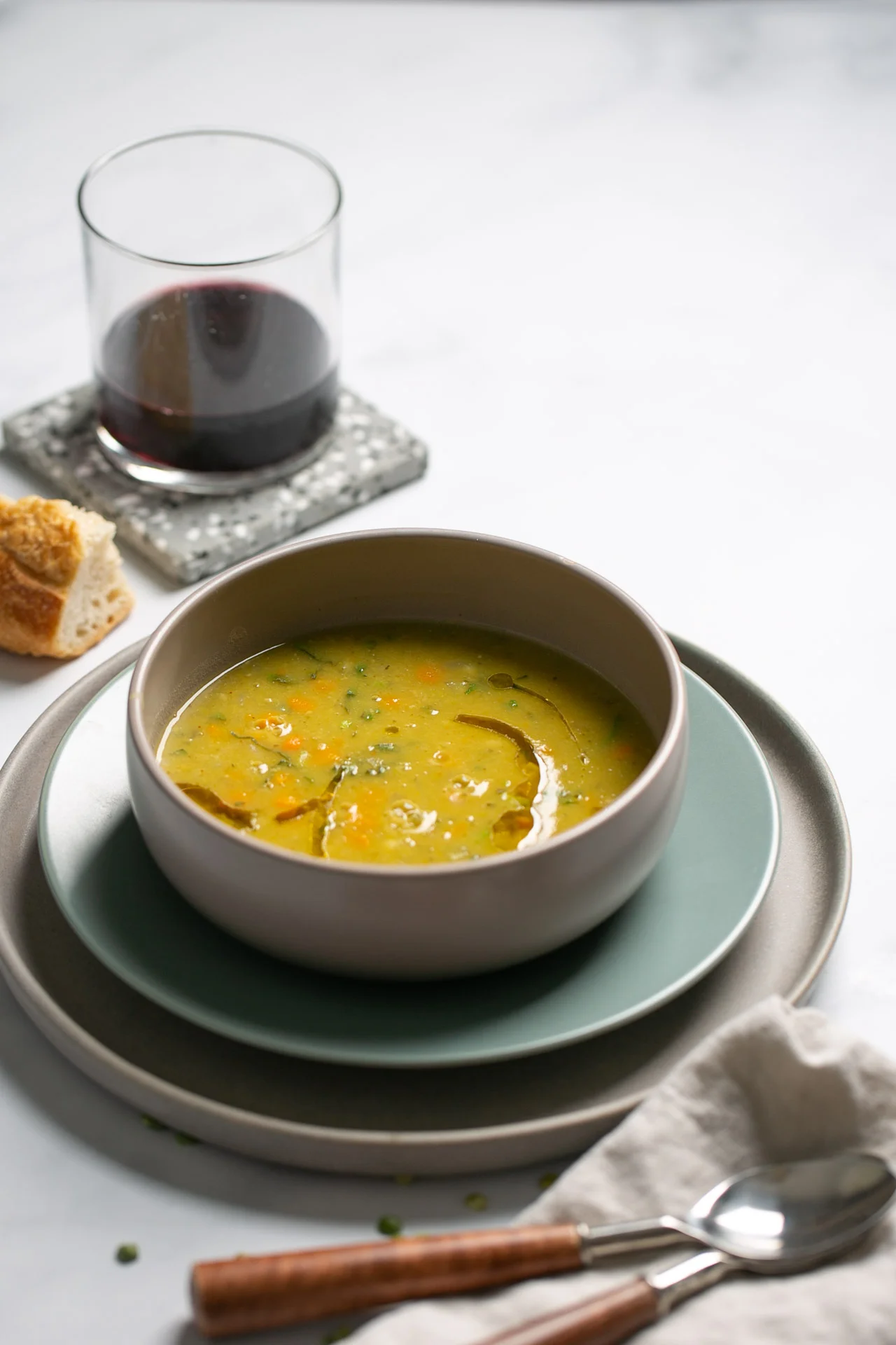 Un cuenco de sopa de verduras de ch&iacute;charo adornada con hierbas se sienta en platos apilados con una servilleta de lino, una cuchara y un tenedor al lado. Al fondo, pan y un vaso de vino tinto sobre un posavasos gris completan la escena sobre una superficie blanca.