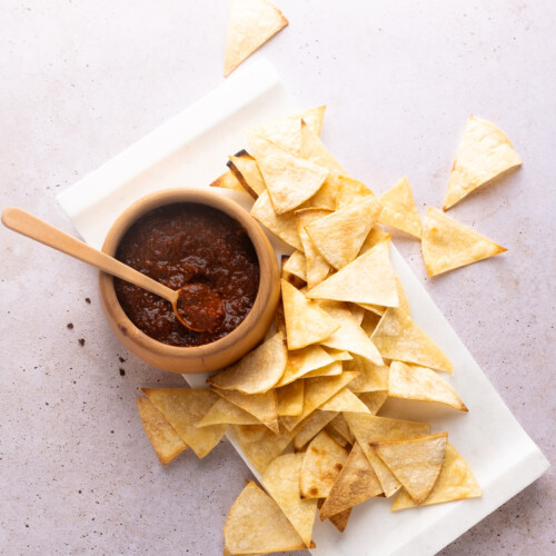 A wooden bowl filled with red salsa and a spoon is placed on a white surface. Surrounding the bowl are scattered triangular corn tortilla chips on a piece of parchment paper. The backdrop is a light, textured stone surface, adding a rustic feel to the composition.