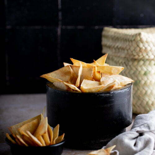 A bowl and a small dish filled with crispy Air Fryer tortilla chips on a dark backdrop.