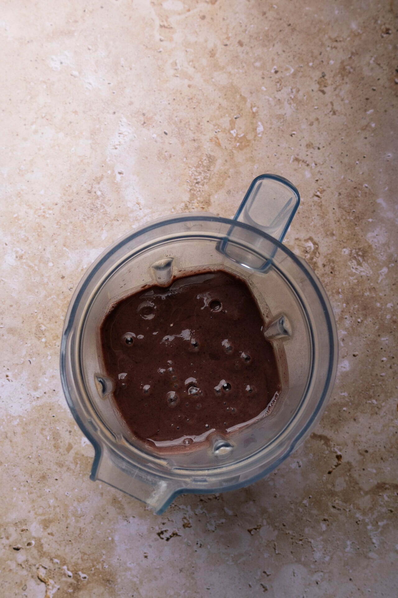Vista desde arriba de una jarra de batidora que contiene un líquido espeso de color marrón oscuro con burbujas en la superficie, posiblemente frijoles puercos o un batido de chocolate, sobre una encimera beige con textura.