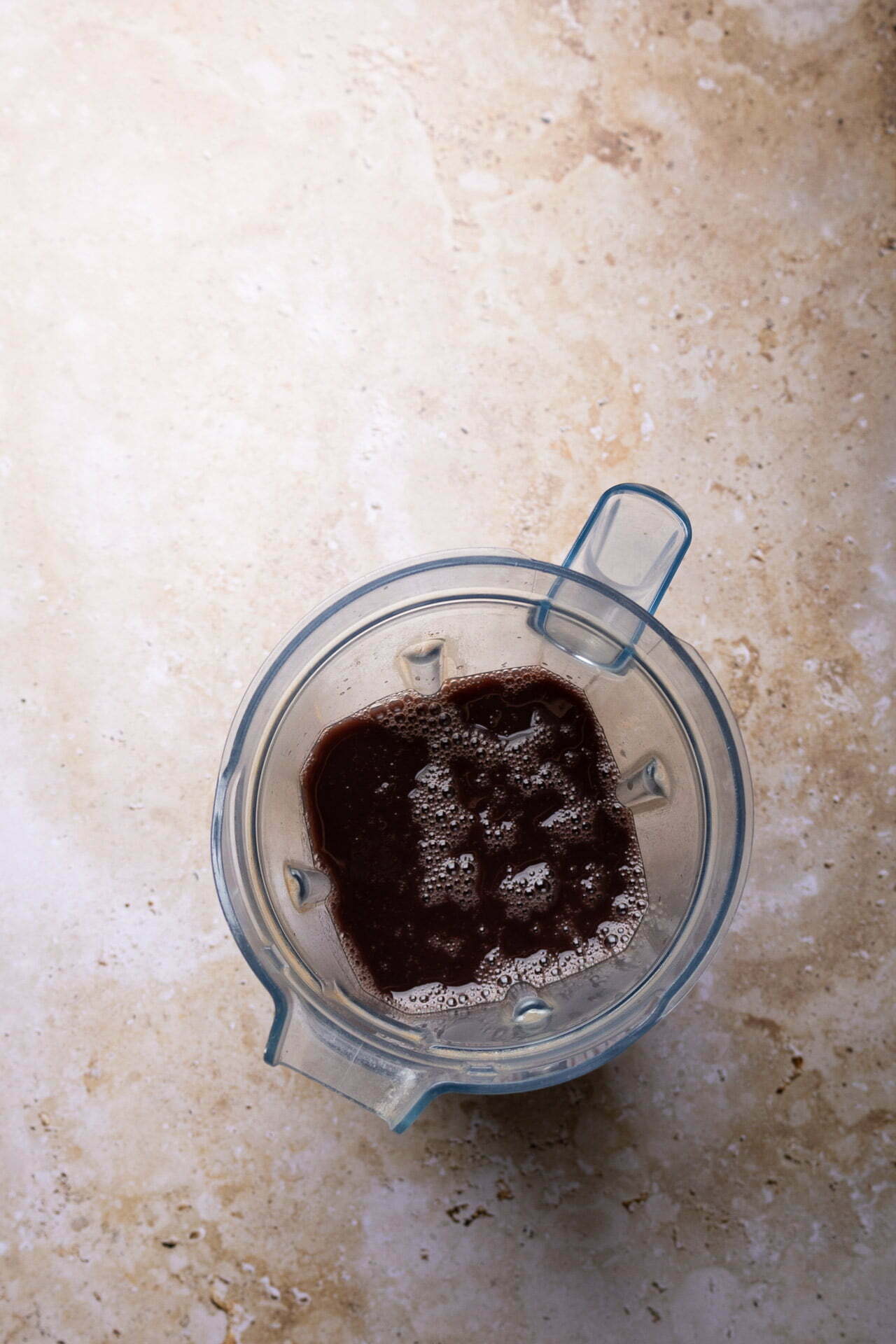 Vista desde arriba de una jarra de batidora que contiene frijoles puercos con una pequeña cantidad de líquido marrón oscuro, burbujas espumosas y trozos de hielo visibles, colocada sobre una encimera de textura beige claro.