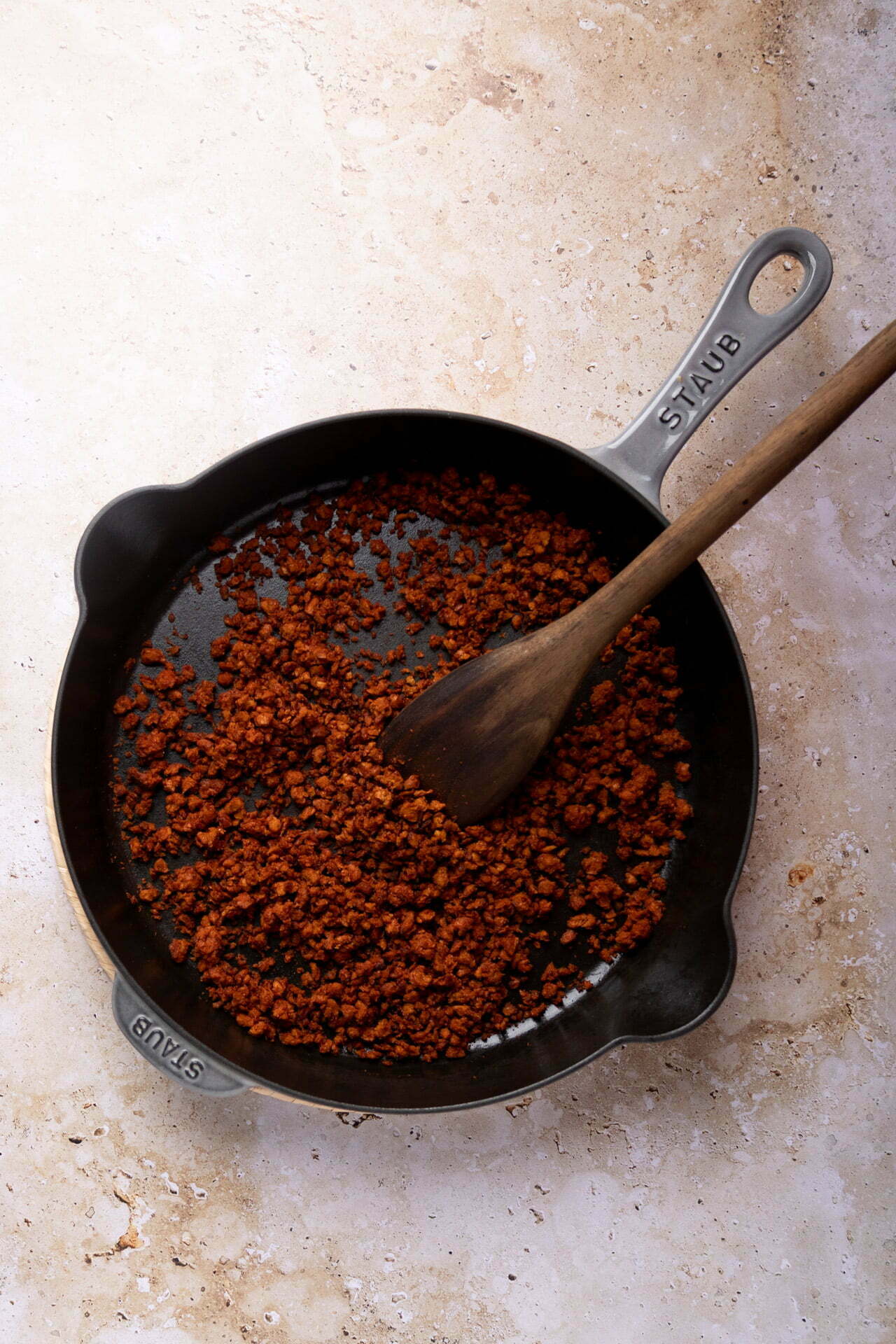 Una sartén de hierro fundido con carne picada dorada y desmenuzada descansa sobre una superficie clara y moteada. Una cuchara de madera descansa dentro de la sartén, revolviendo la carne cocida-perfectamente preparada para agregar a los frijoles puercos u otros platos sustanciosos.