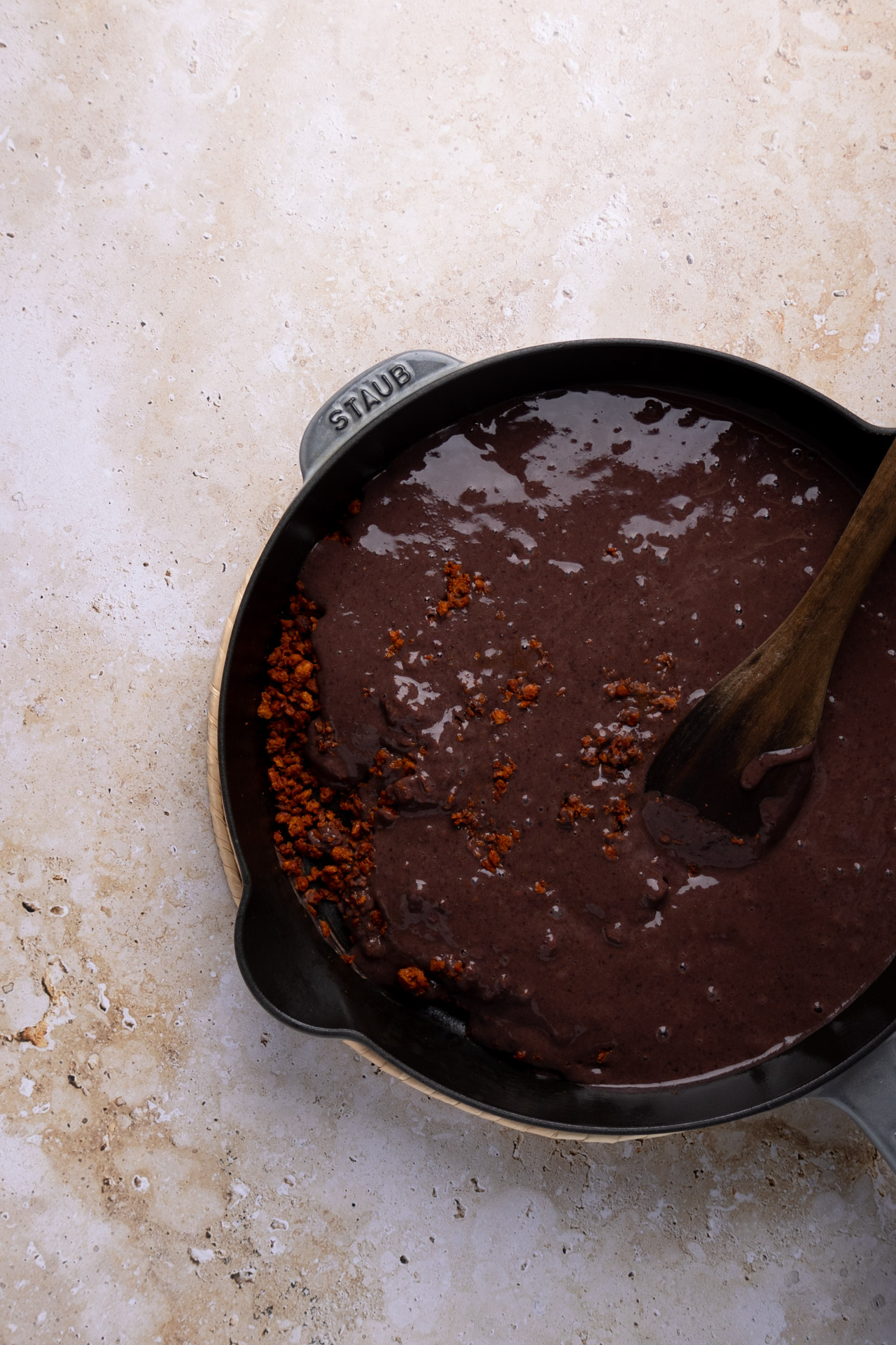 Una sartén de hierro fundido negro llena de oscuros y gruesos frijoles puercos y chorizo cocido de color marrón rojizo se asienta sobre una superficie de piedra de color beige claro. Una cuchara de madera se apoya en la sartén, mezclando parcialmente los ingredientes. El mango de la sartén lleva grabado "STAUB".