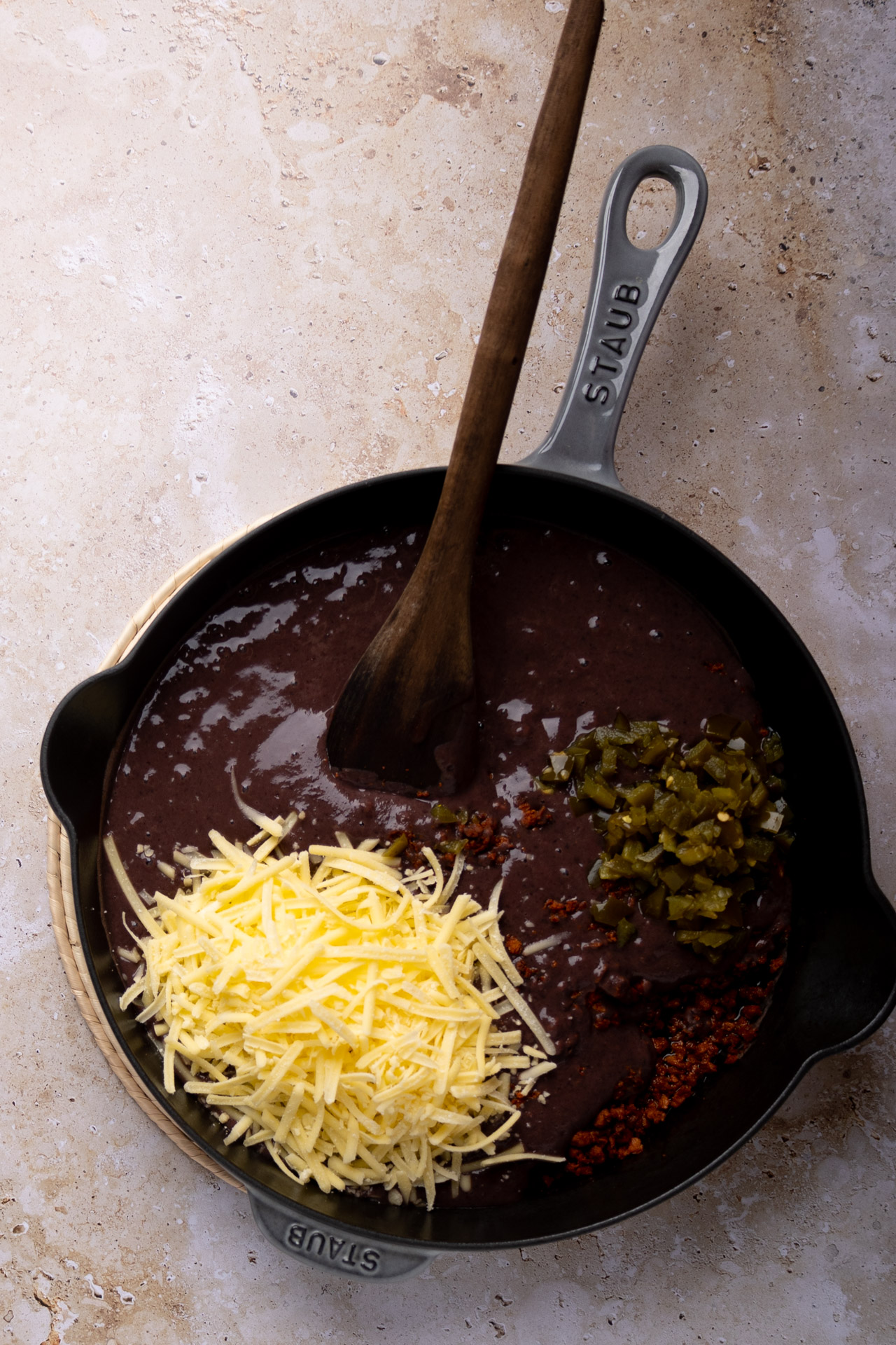 Una sartén de hierro fundido llena de cremosos frijoles puercos descansa sobre una superficie de piedra clara. Dentro hay tres ingredientes: queso cheddar rallado, chiles verdes picados y un montón de hojuelas de chile rojo. Dentro hay una cuchara de madera.