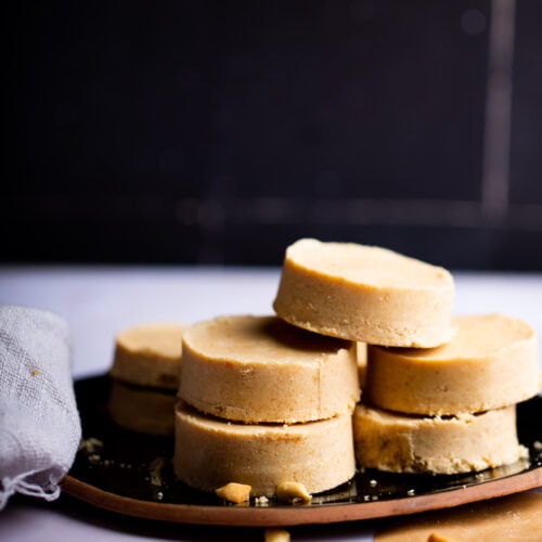 A stack of five round, beige cookies is on a dark plate against a black background. They resemble crumbly and rich mazapán, with cookie crumbs scattered on the white surface. To the side, a light gray cloth adds texture to the scene.
