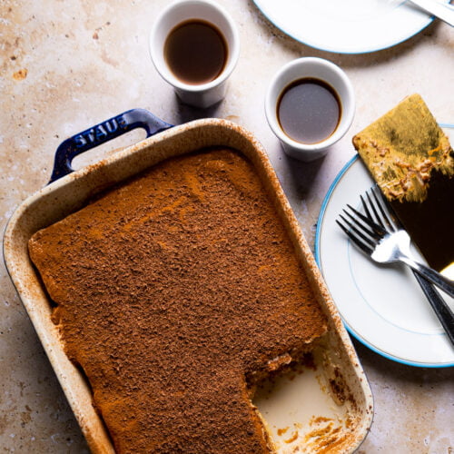 Sobre una superficie de mármol claro, espolvoreada con cacao en polvo, reposa un plato de tiramisú vegano al que le falta una pieza. Dos tazas blancas llenas de café acompañan a dos platos, uno de los cuales contiene una rodaja de triamisu sin soletas. Los tenedores están colocados elegantemente sobre los platos.