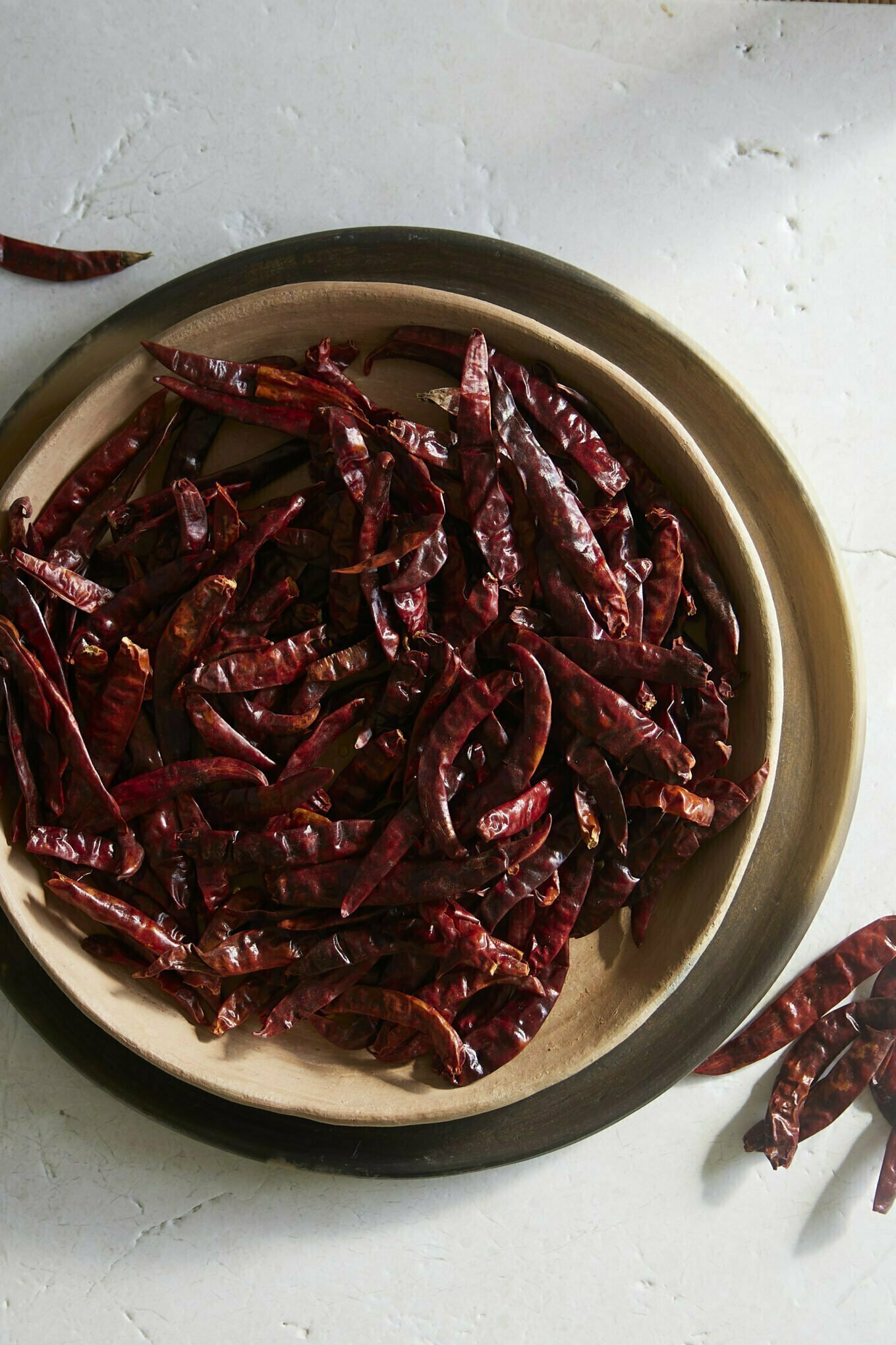 A wooden bowl filled with numerous dried Chile de Arbol chilies sits on a light-colored, textured surface. Several fiery Mexican chiles have spilled out onto the table, their vibrant hues and shapes highlighted by soft shadows.