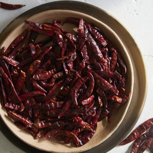 Un cuenco beige lleno de chiles rojos secos está colocado en una bandeja redonda de madera sobre una superficie clara y texturizada. Fuera del cuenco hay esparcidos varios chiles y totopos, que añaden un toque rústico a la escena mientras las suaves sombras juegan sobre ella.