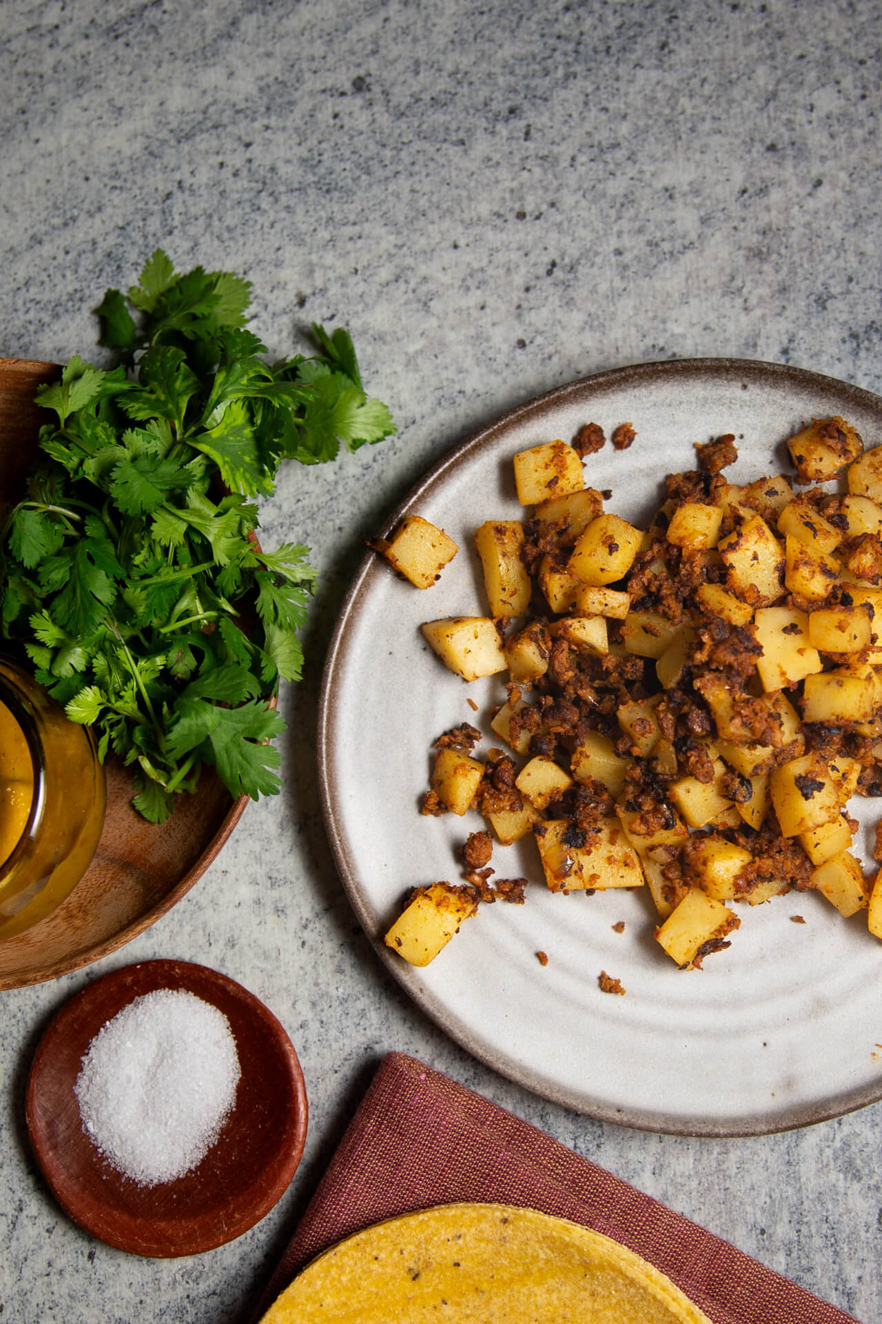 Un plato de patatas cortadas en cubitos y sazonadas &mdash;perfectas para unas &laquo;papas con chorizo&raquo; veganas&mdash; descansa sobre una superficie gris junto a un cuenco de madera con cilantro fresco, un platito con sal, un frasco de aceite y una tortilla de ma&iacute;z amarilla que se vislumbra parcialmente en el fondo. Debajo del plato hay una servilleta roja, que crea el ambiente perfecto para esta deliciosa receta vegana.