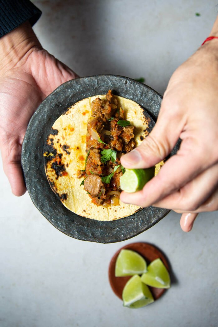 Hombre sosteniendo plato oscuro con bistec a la mexicana sobre una tortilla de maíz y un poco de lima siendo exprimida