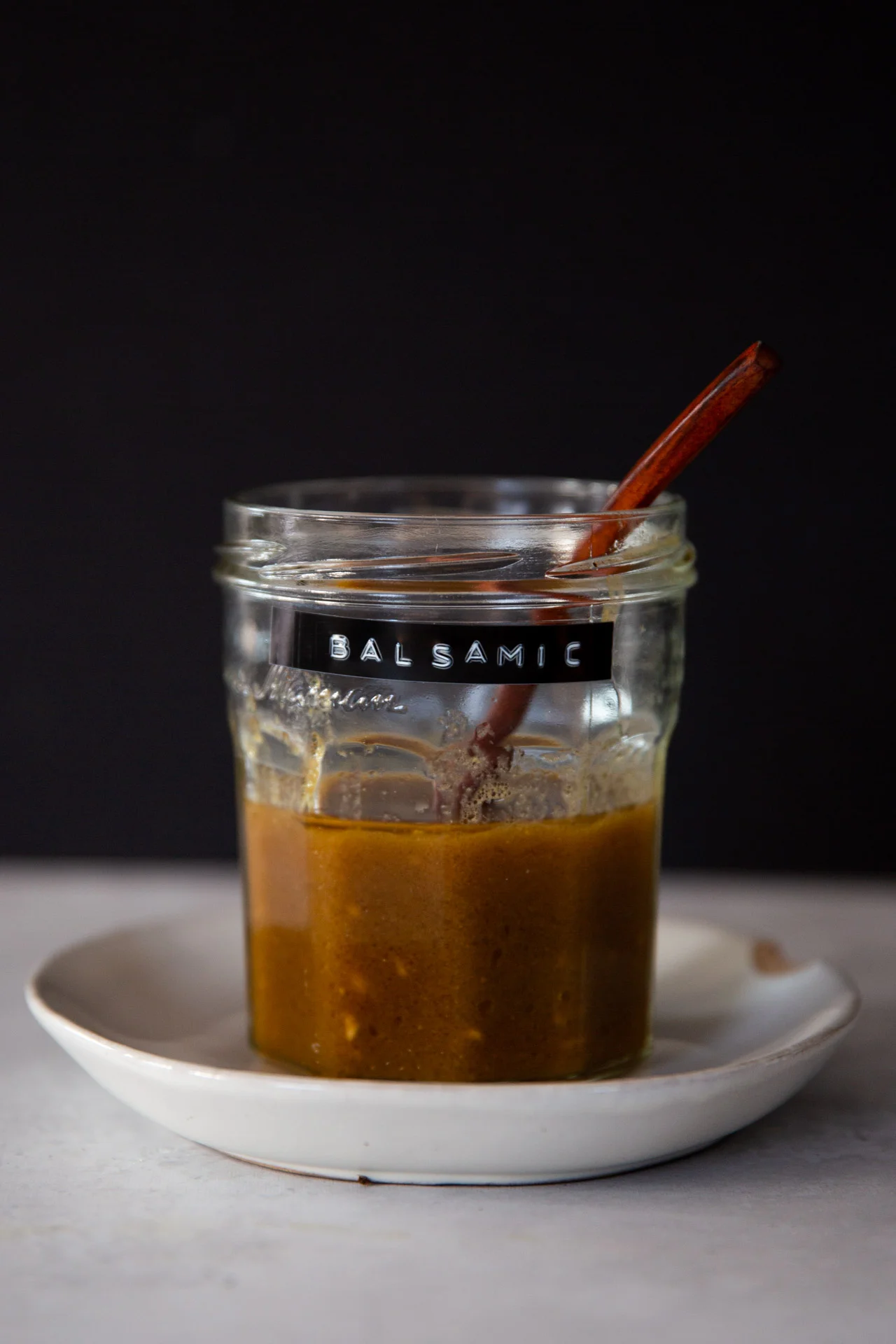 Sobre un plato blanco hay un tarro de cristal transparente con la etiqueta "BALSAMIC" parcialmente lleno de Vinagreta Bals&aacute;mica marr&oacute;n hecha en casa. Dentro del tarro hay una cuchara de madera marr&oacute;n. El fondo oscuro contrasta con el tarro y el plato.