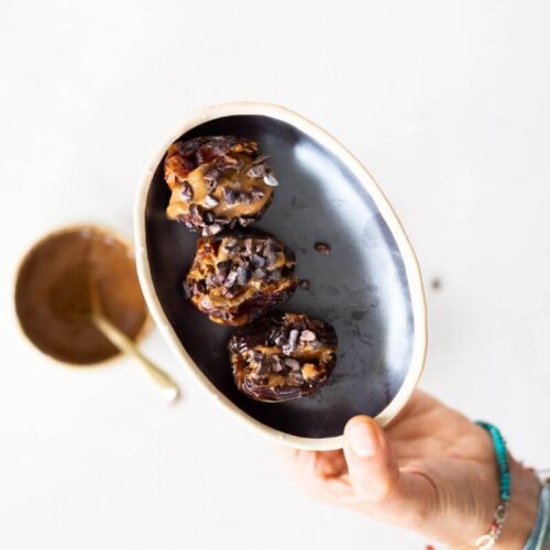 A hand wearing colorful bracelets holds a small, oval black plate with three almond butter stuffed dates topped with chocolate chips. In the background, there is a blurry gold bowl with a spoon on a white surface.