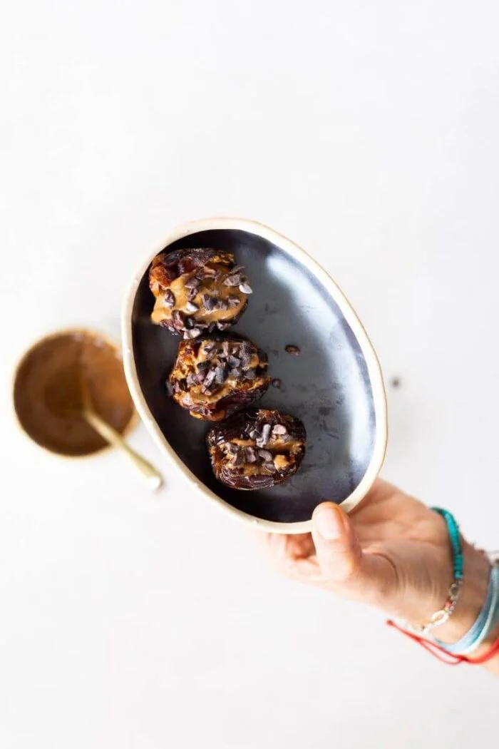 A hand wearing colorful bracelets holds a small, oval black plate with three almond butter stuffed dates topped with chocolate chips. In the background, there is a blurry gold bowl with a spoon on a white surface.