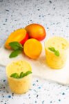 Two glasses of vegano mango lassi garnished with mint leaves rest on a white marble board with ice. Nearby, whole and halved ripe mangoes contrast beautifully against the speckled white surface, highlighting the vibrant orange hues of both the mangoes and lassi.