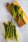 Varios tallos de espárragos verdes frescos se disponen sobre una tabla de cortar de madera con un lazo de cuero y sobre un plato ovalado blanco, ambos colocados sobre una superficie moteada de gris claro. Perfectos para su próxima receta utilizando el sartén. Las puntas de los espárragos son puntiagudas y vibrantes.