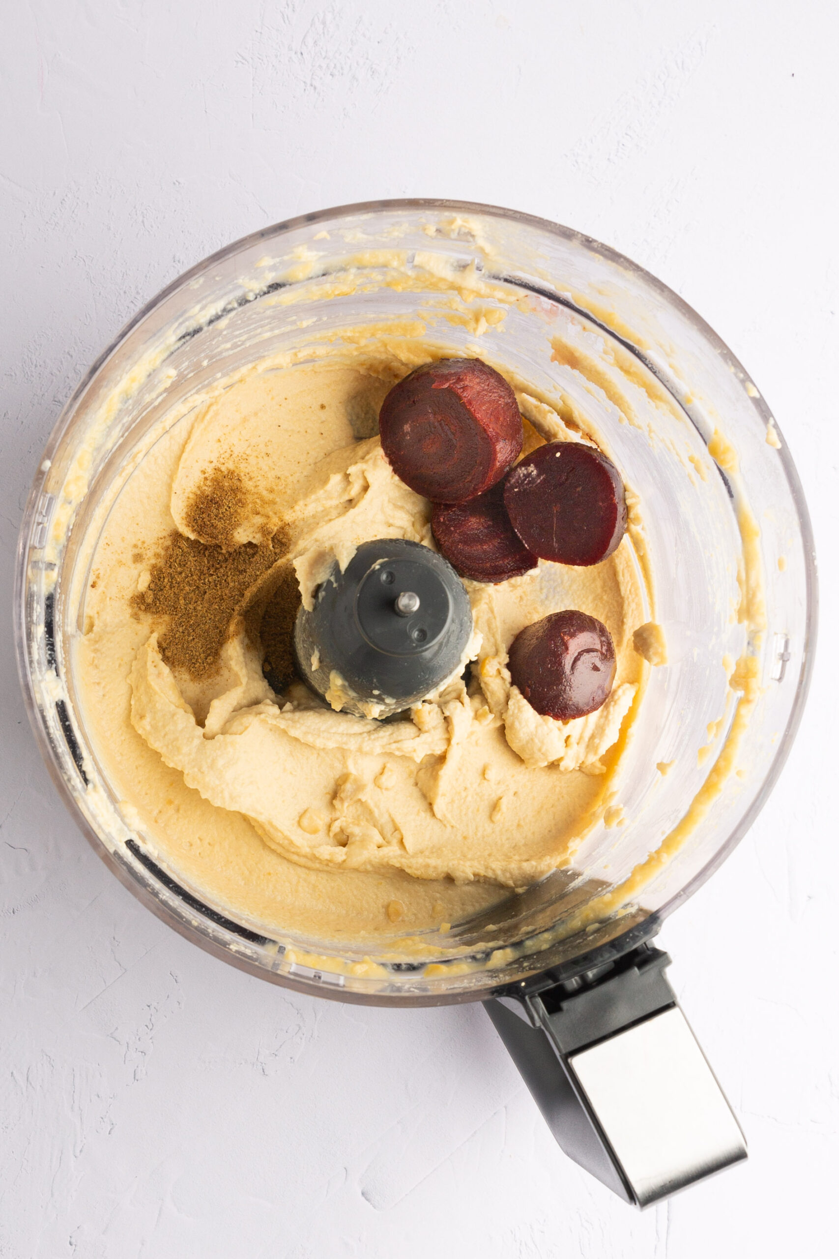 Hummus, betabeles asados y comino en el tazón de un procesador de alimentos, listos para licuar.