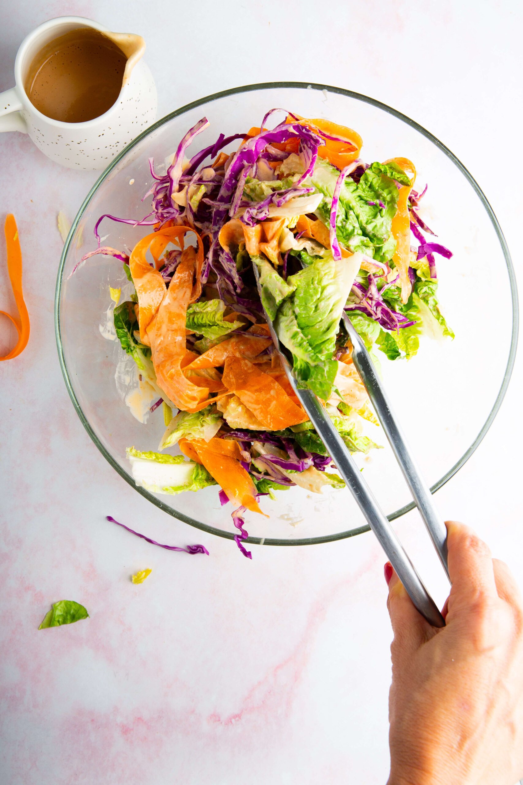 A hand uses metal tongs to mix an ensalada crujiente with lettuce, purple cabbage, and carrot ribbons in a glass bowl. A small white jug of aderezo de cashew sits nearby on a light pink surface. A few pieces of salad are scattered on the table.