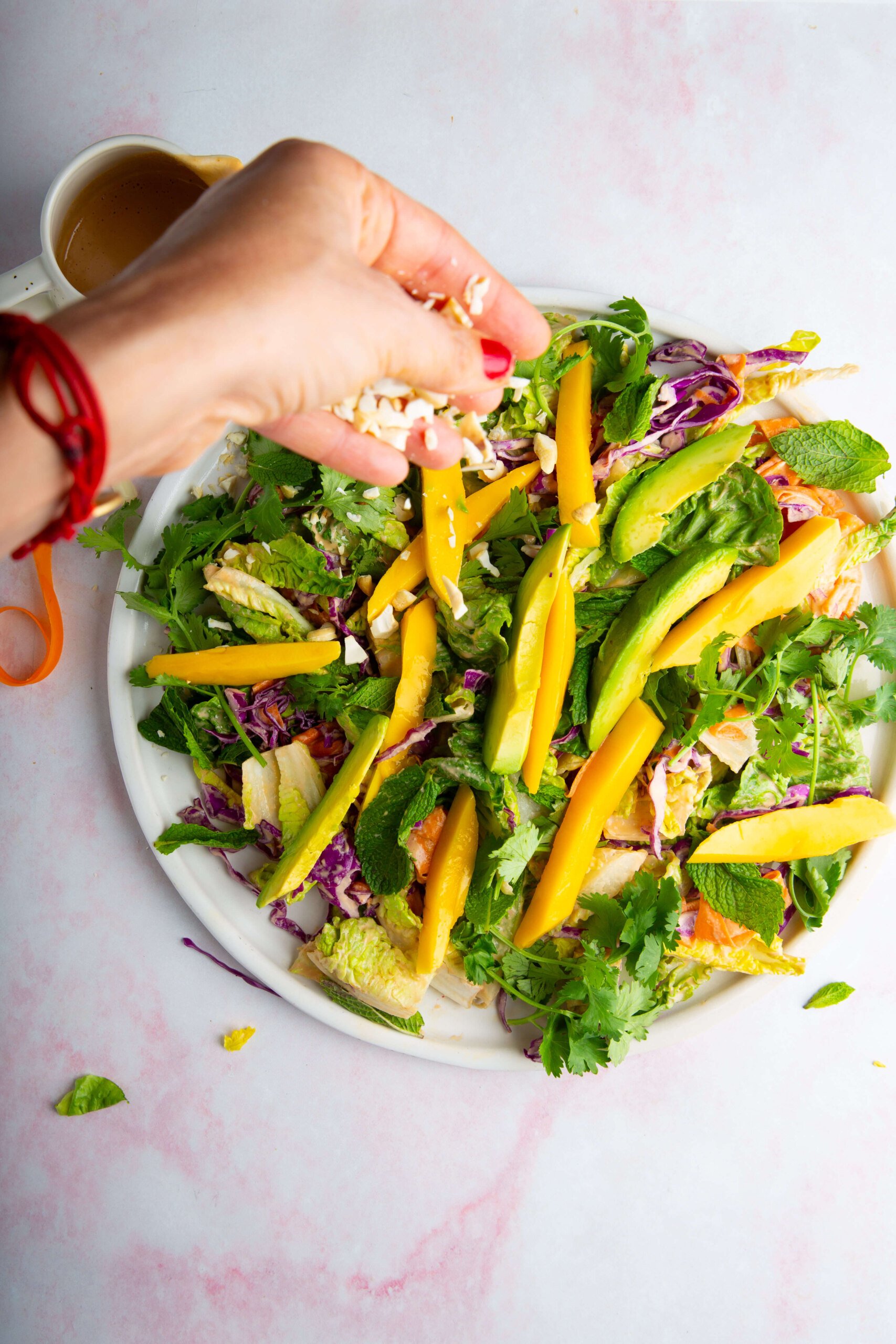 A hand with red nail polish sprinkles shredded coconut over an ensalada crujiente with mango slices, avocado, fresh herbs, shredded cabbage, and greens on a white plate. A small cup of aderezo de cashew sits nearby on a light pink marbled surface.