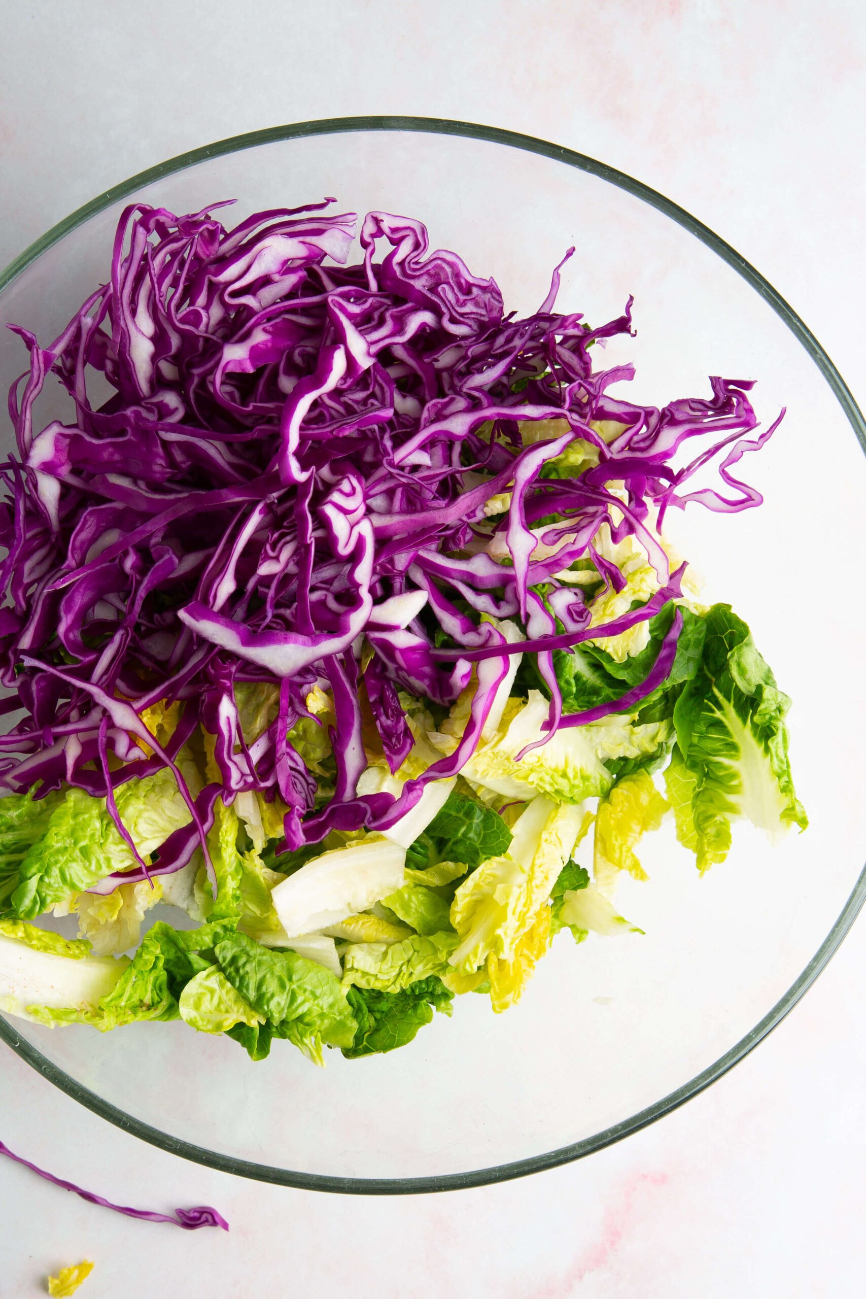 A clear glass bowl filled with fresh, shredded purple cabbage and chopped green romaine lettuce sits on a white surface, creating an ensalada crujiente. The vibrant vegetables await a drizzle of aderezo de cashew for extra flavor.