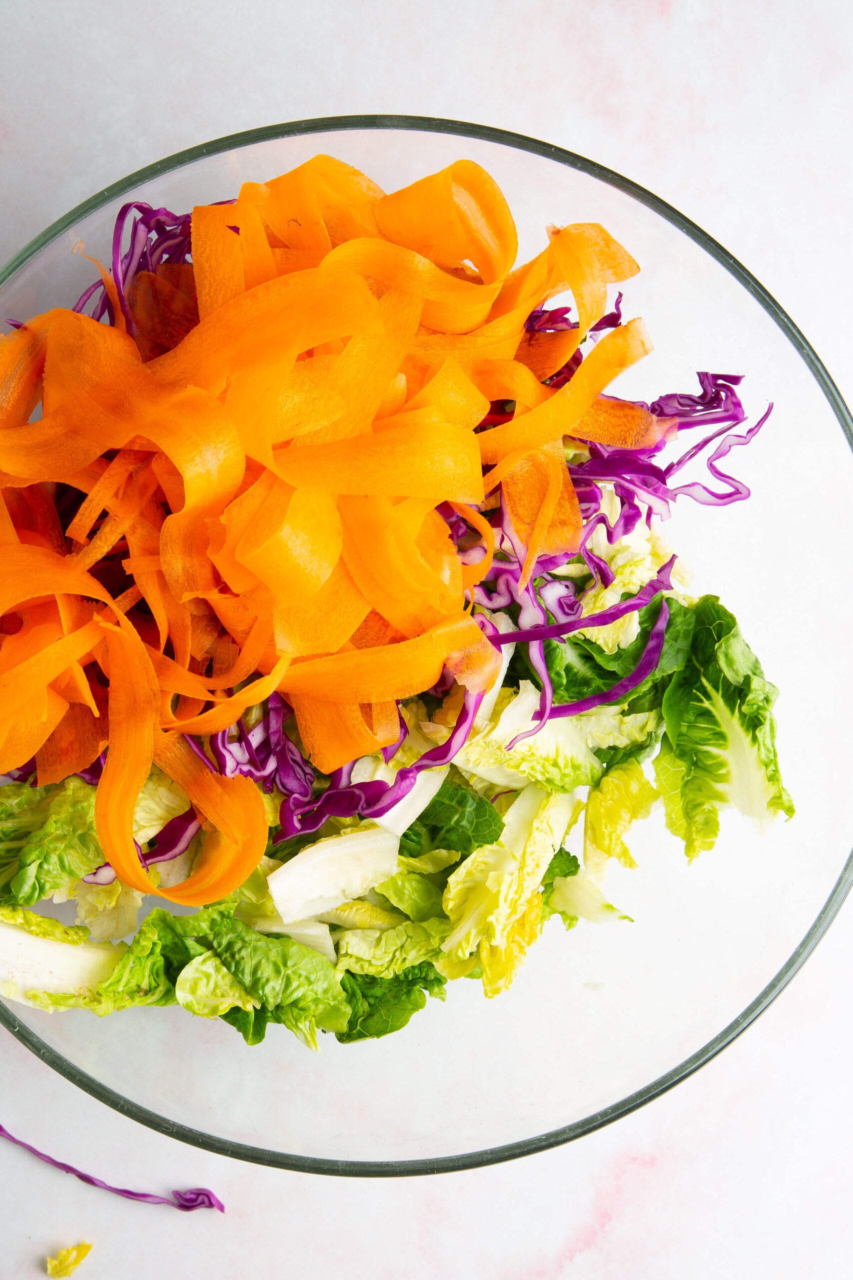A clear glass bowl filled with ensalada crujiente ingredients—green romaine lettuce, thinly shredded purple cabbage, and wide orange carrot ribbons—layered vibrantly on a light pink background. Perfect with a drizzle of aderezo de cashew.