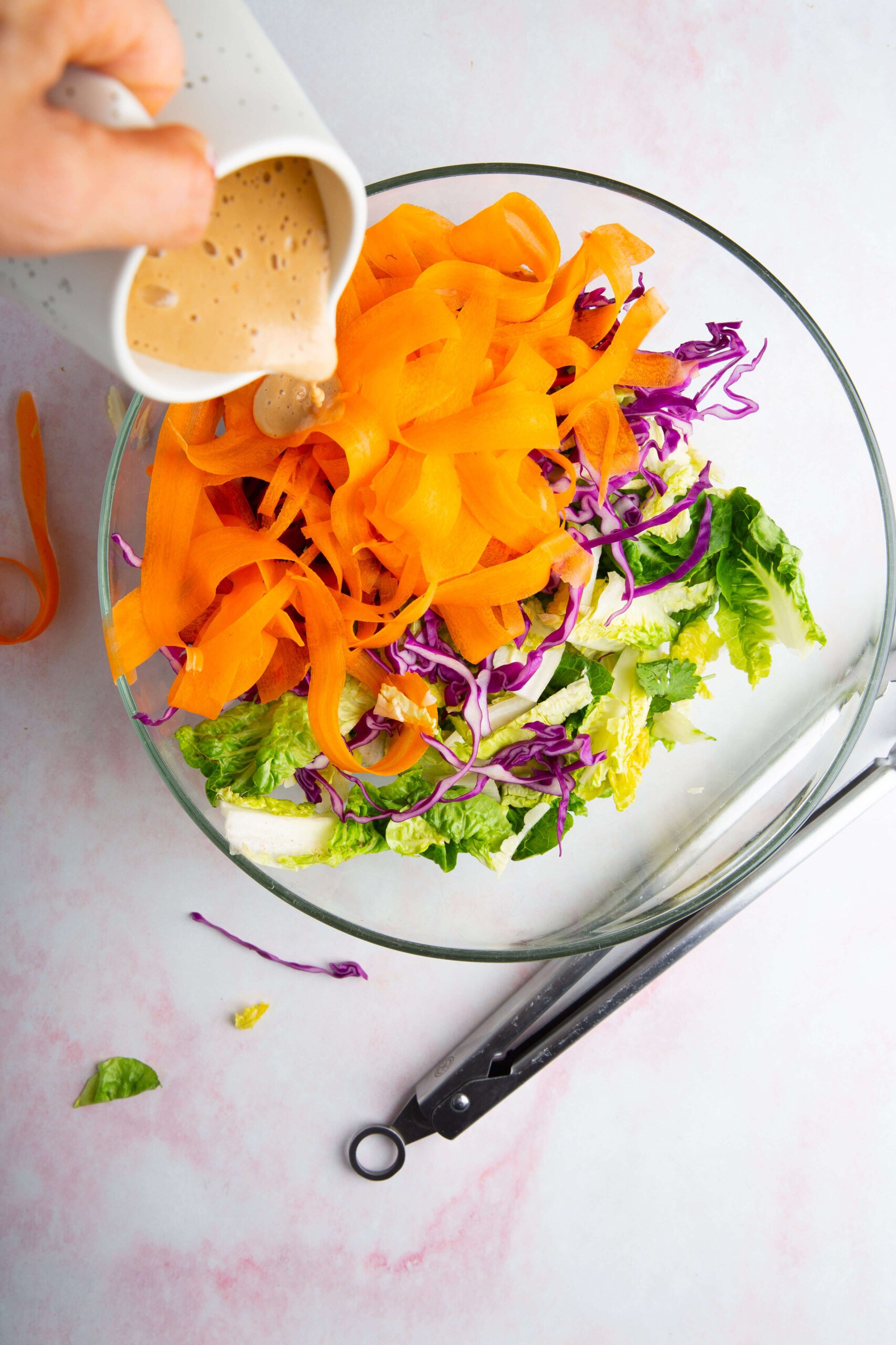 A hand pours creamy aderezo de cashew from a white pitcher onto an ensalada crujiente of ribboned carrots, shredded purple cabbage, and lettuce in a glass bowl. Metal tongs rest nearby on a light pink marbled surface.