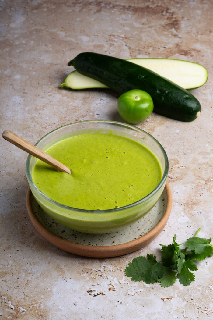A glass bowl with smooth green zucchini salsa and a wooden spoon sits on a plate. Behind it are two zucchinis, one sliced, a green tomatillo, and cilantro leaves on a beige textured surface, hinting at fresh salsa taquera ingredients.