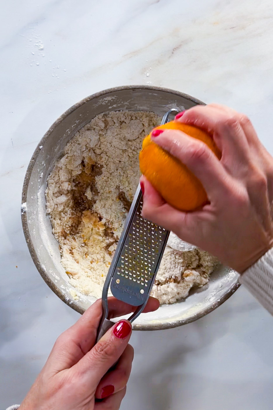 Una persona con las uñas pintadas de rojo ralla ralladura de naranja sobre harina e ingredientes para hornear, preparando galletas de Día de los Muertos sobre una encimera blanca, perfecta para recetas mexicanas y crear galletas festivas y decoradas.