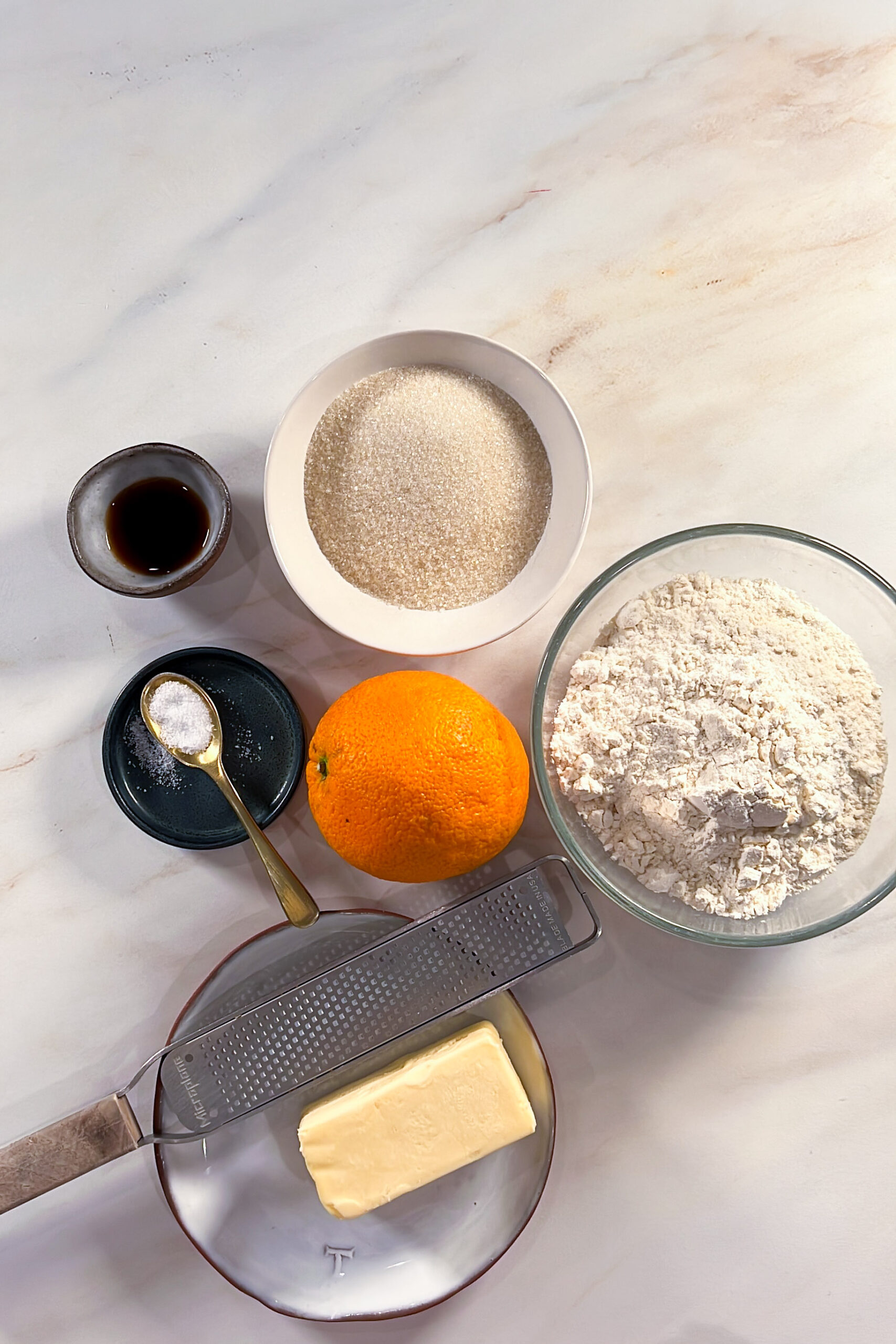 Coloque los ingredientes para las galletas de Día de los Muertos sobre una superficie clara: azúcar, harina, extracto de vainilla, sal con una cuchara, una naranja entera y mantequilla en un plato con un rallador de metal.